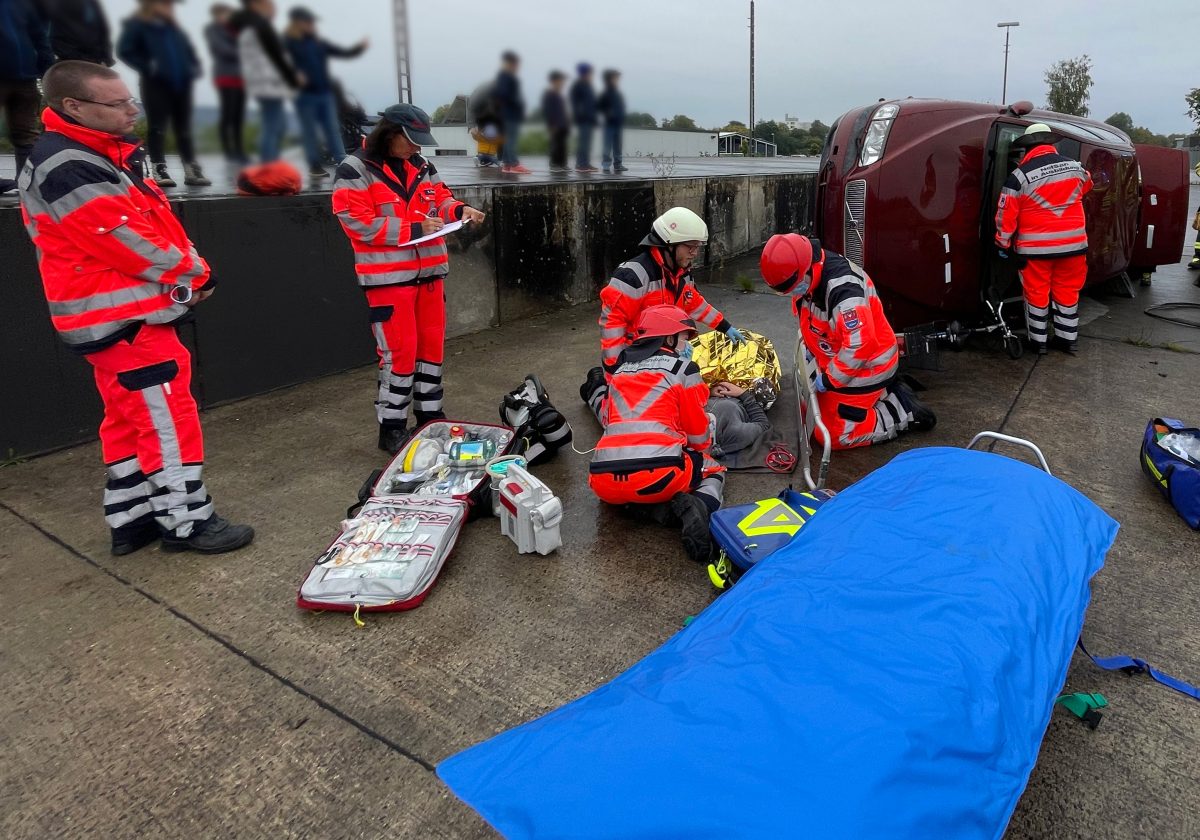 Rettungsfachpersonal aus sechs Personen an einem nachgestellten Unfallort mit einem umgekehrten Auto und einer am Boden liegenden Person