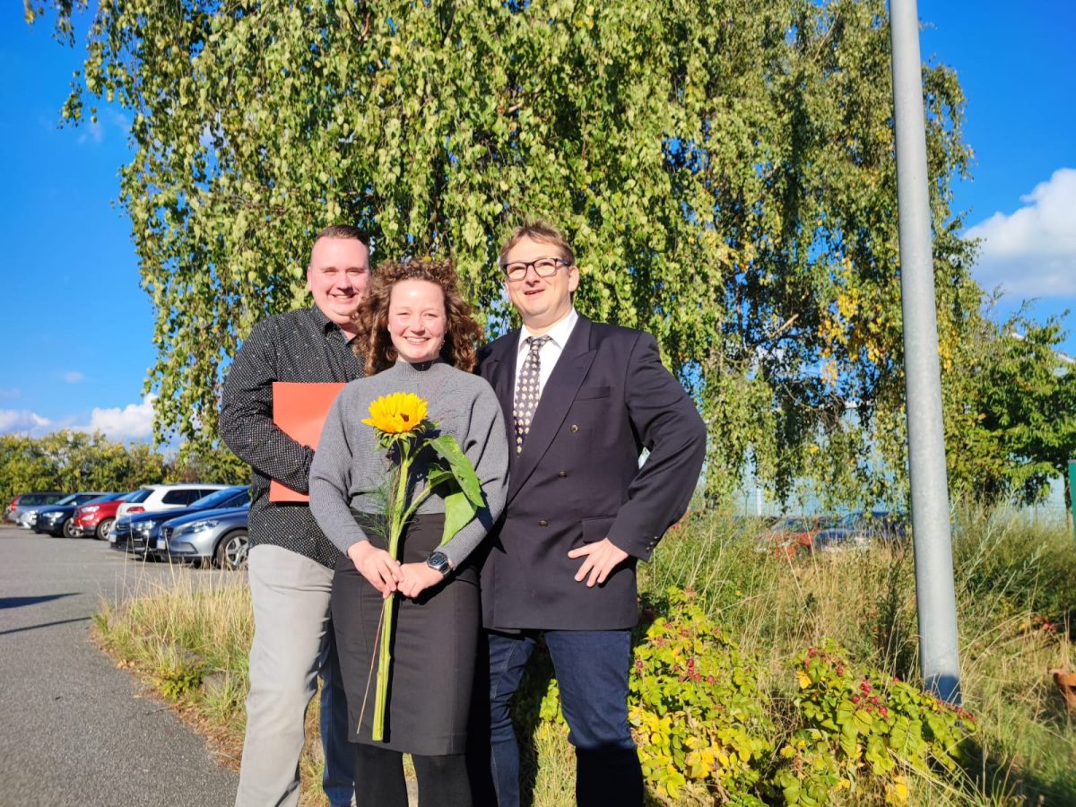 Drei Personen lächeln in die Kamera mit einer Blume und einem Zeugnis